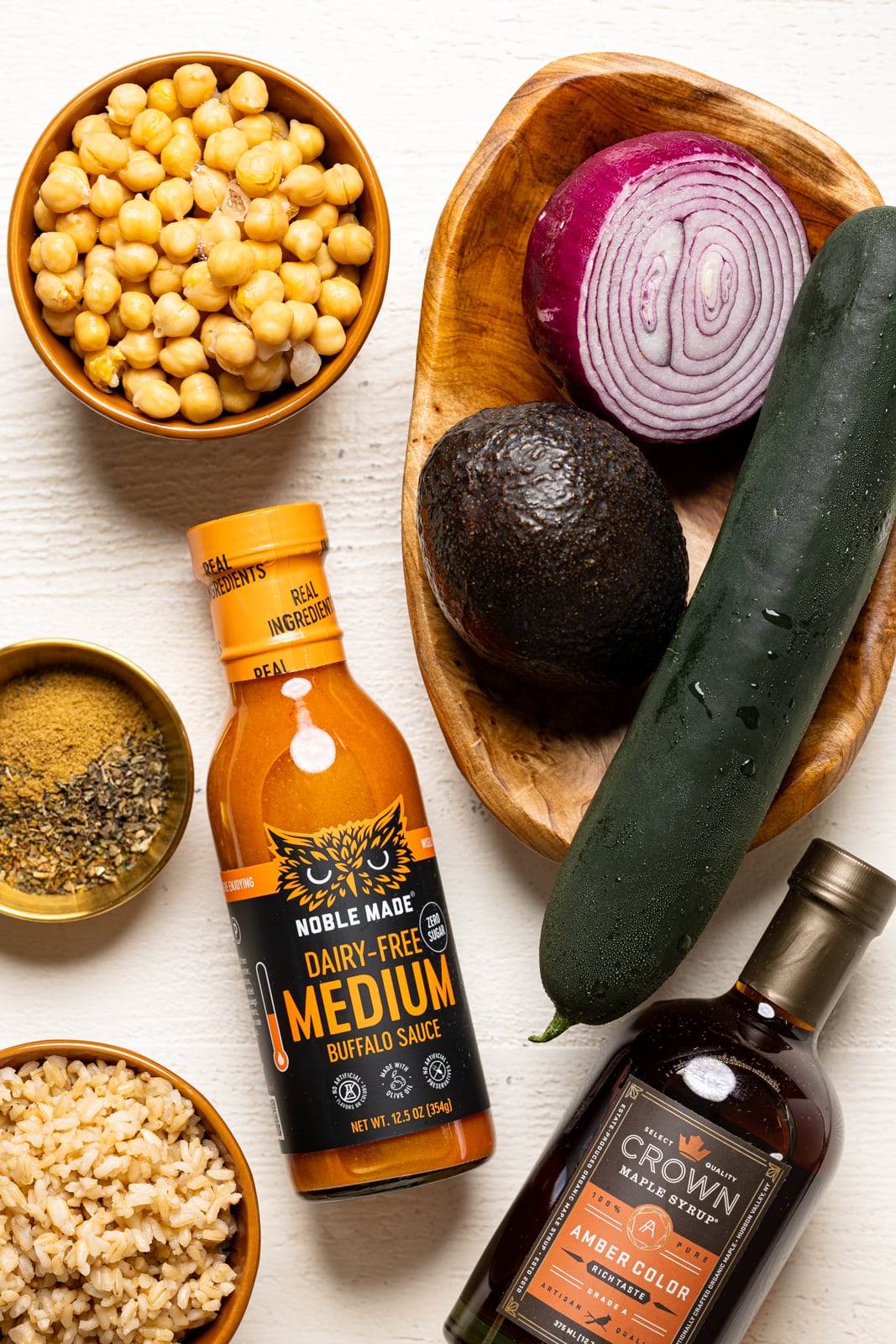 Ingredients on a white wood table including chickpeas, buffalo sauce, maple syrup, brown rice, herbs and seasonings, and veggies.