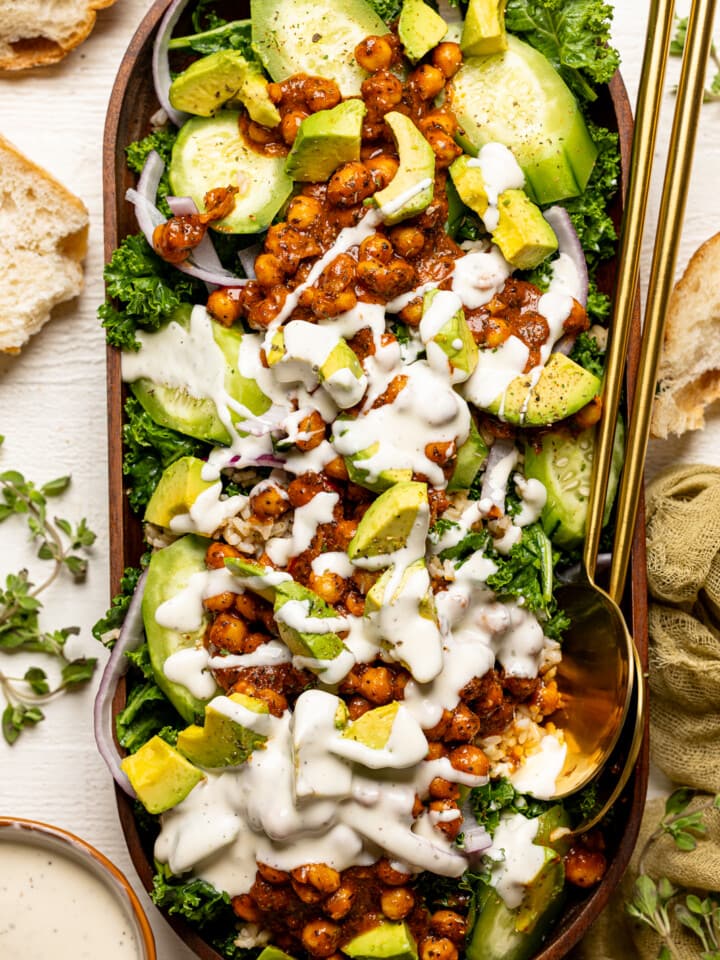 Fully loaded Kale salad with white dressing in a large wooden serving dish with two gold spoons.