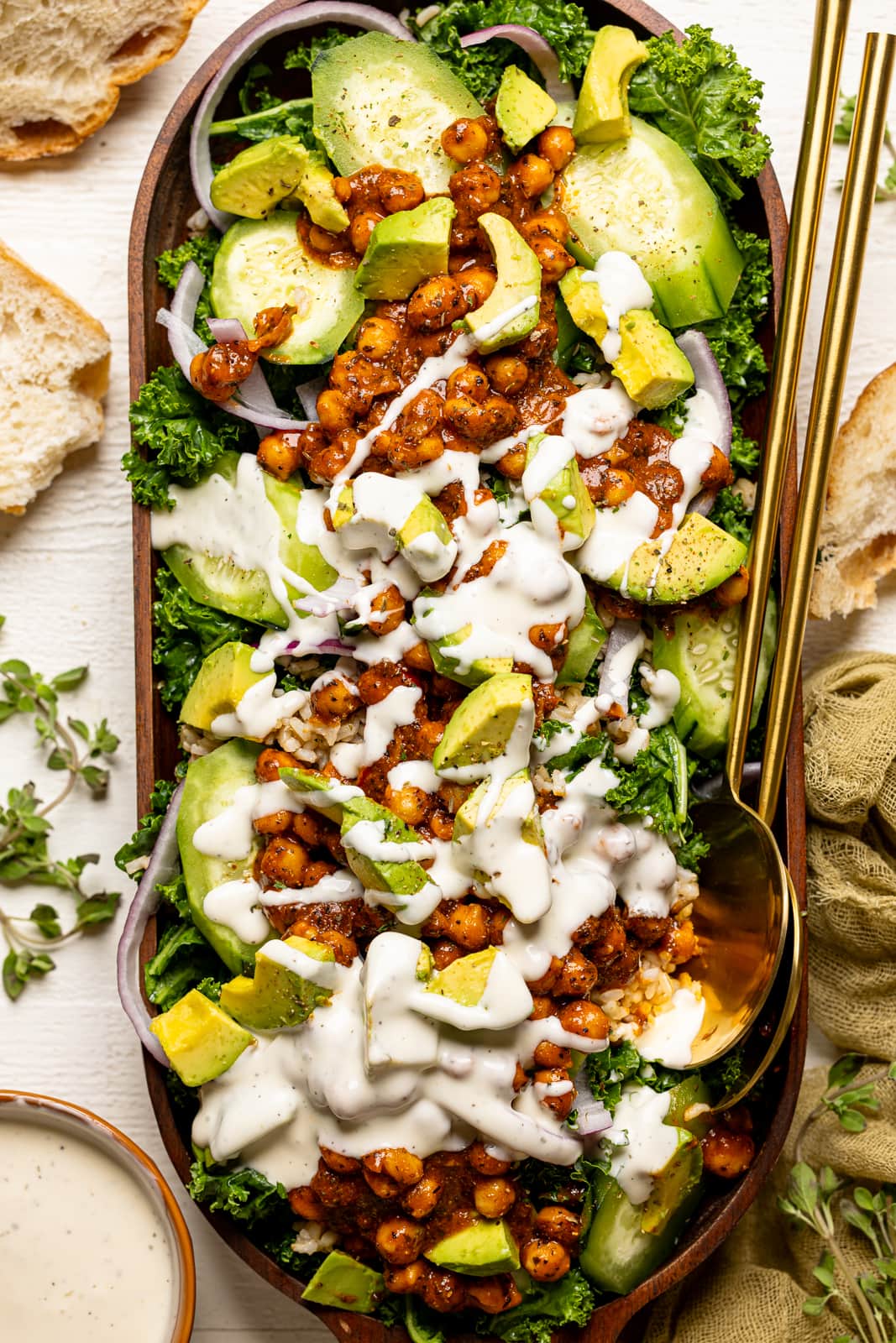 Fully loaded Kale salad with white dressing in a large wooden serving dish with two gold spoons.