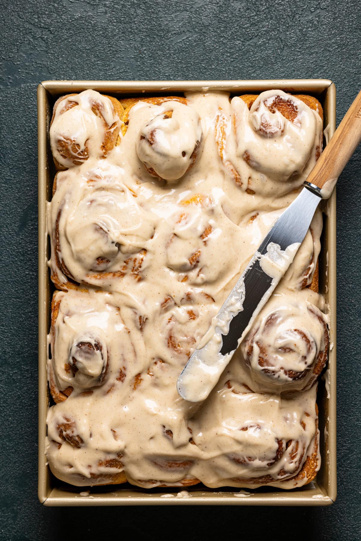 Frosted cinnamon rolls in a baking dish with a spatula. 