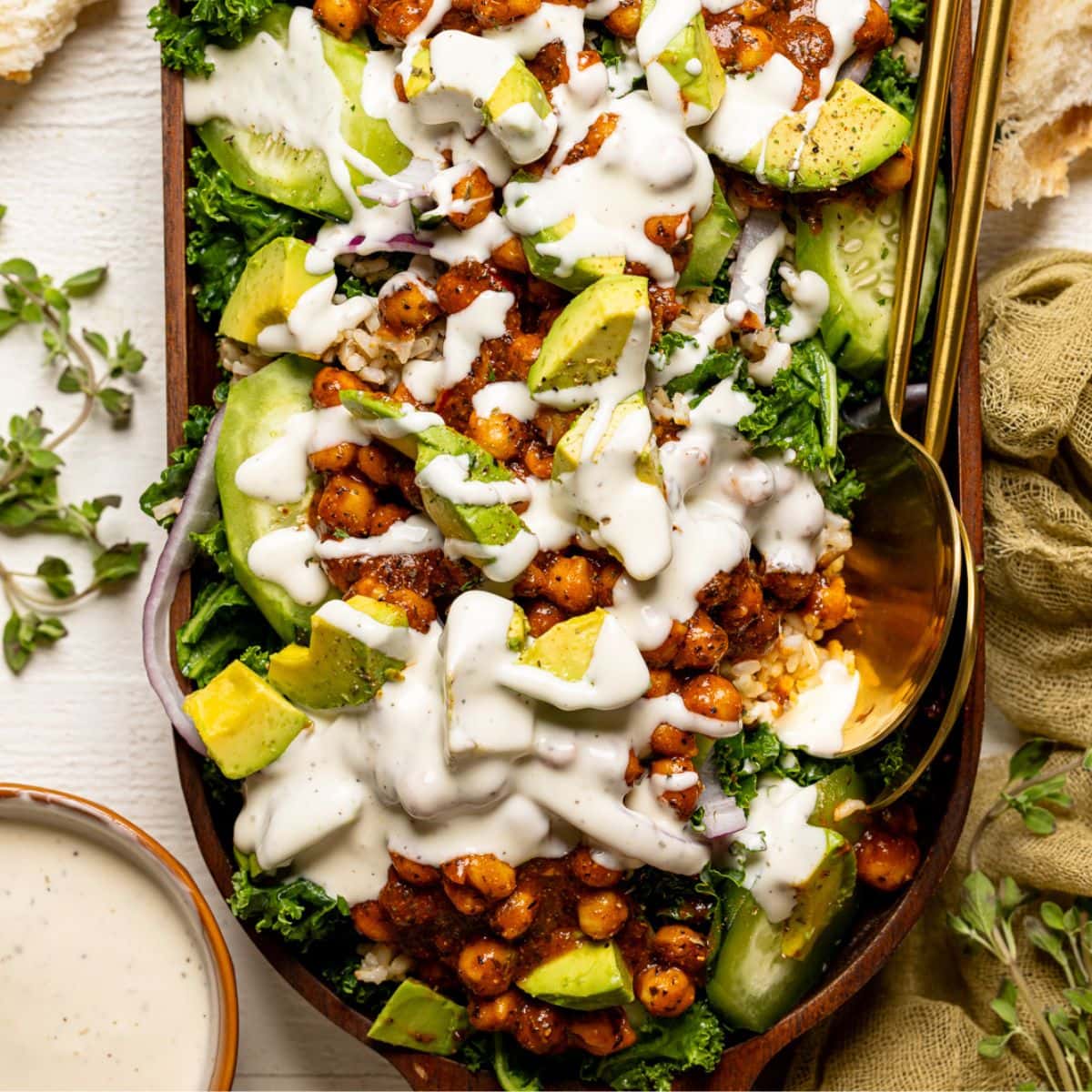 Fully loaded Kale salad with white dressing in a large wooden serving dish with two gold spoons.