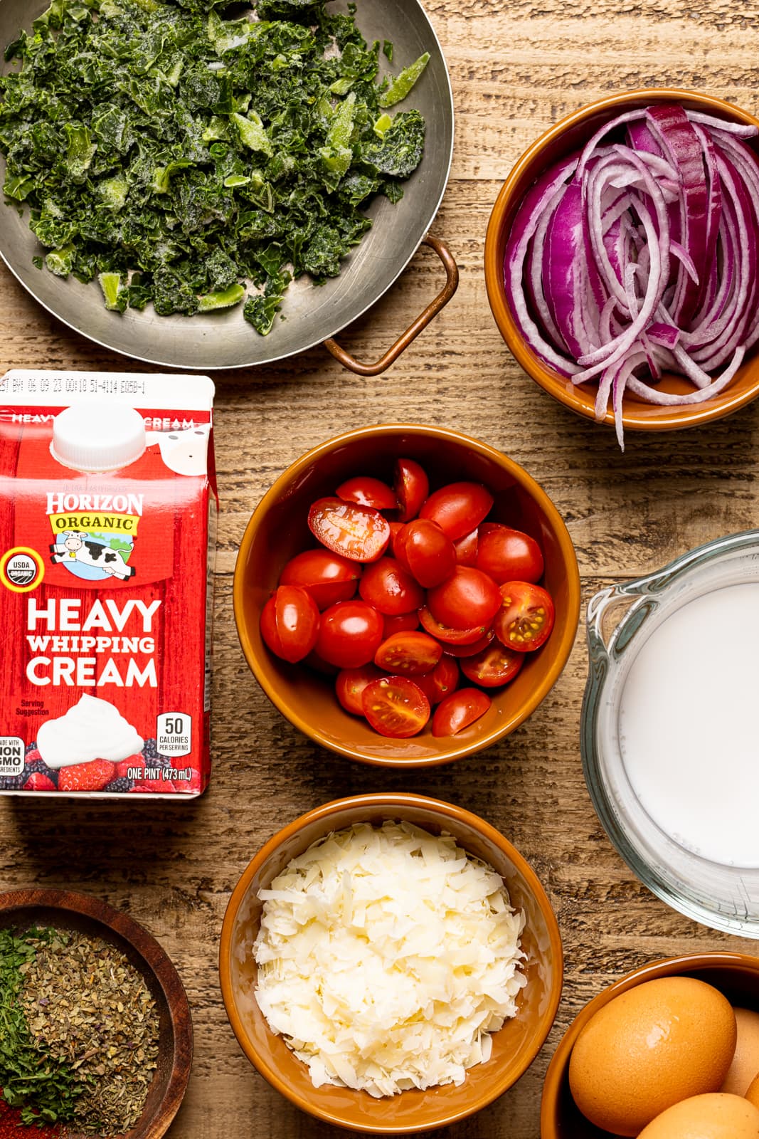 Ingredients on a brown wood table including heavy cream, tomatoes, onions, kale, milk, and grated parmesan cheese.