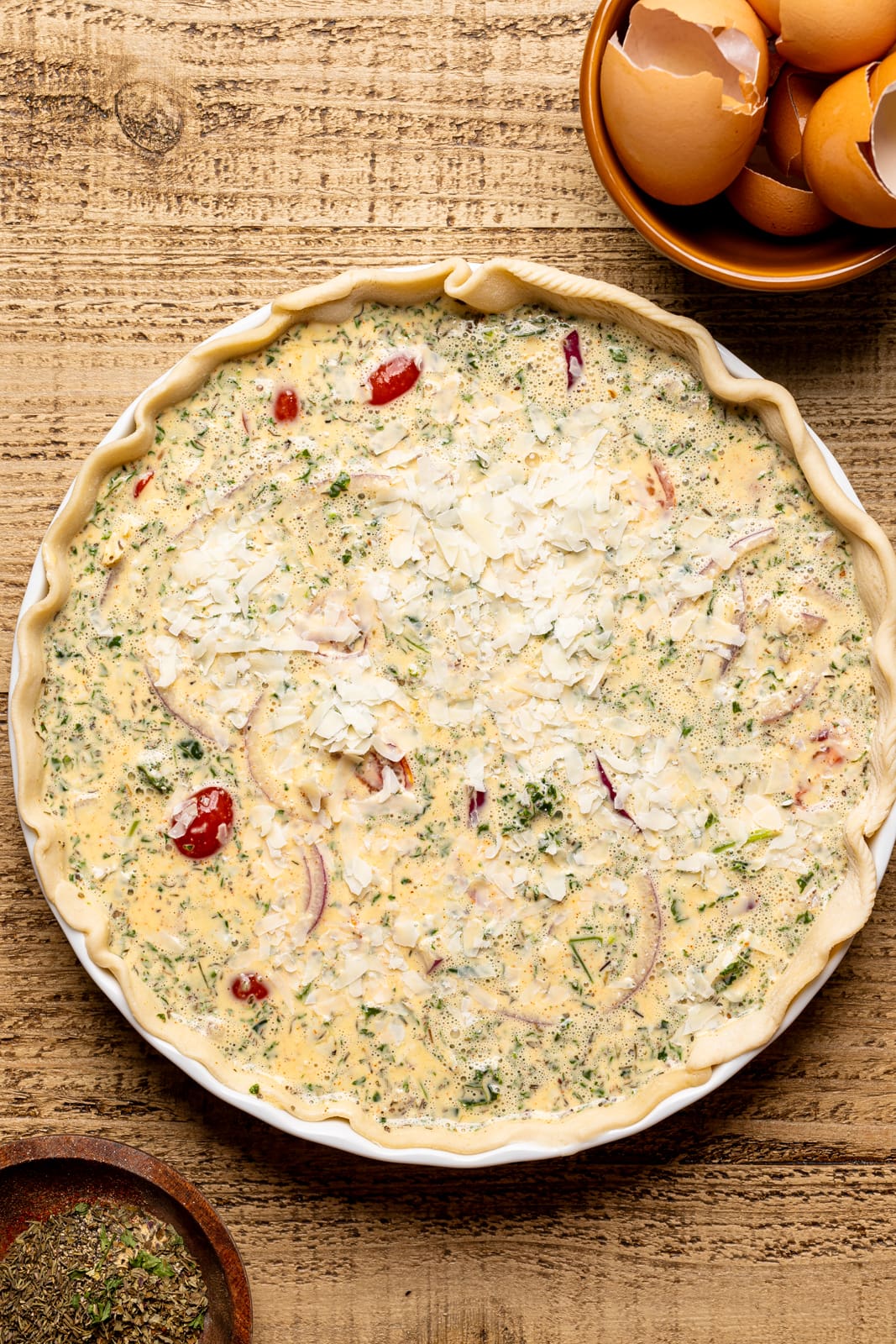 Quiche mixture poured unto crust in a baking dish on a brown wood table with herbs + seasonings.
