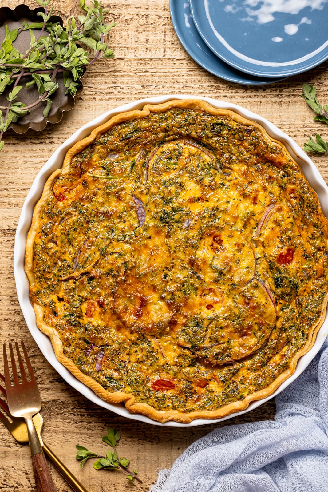 Baked quiche in a baking dish on a brown wood table with herbs + seasonings and a blue napkin.