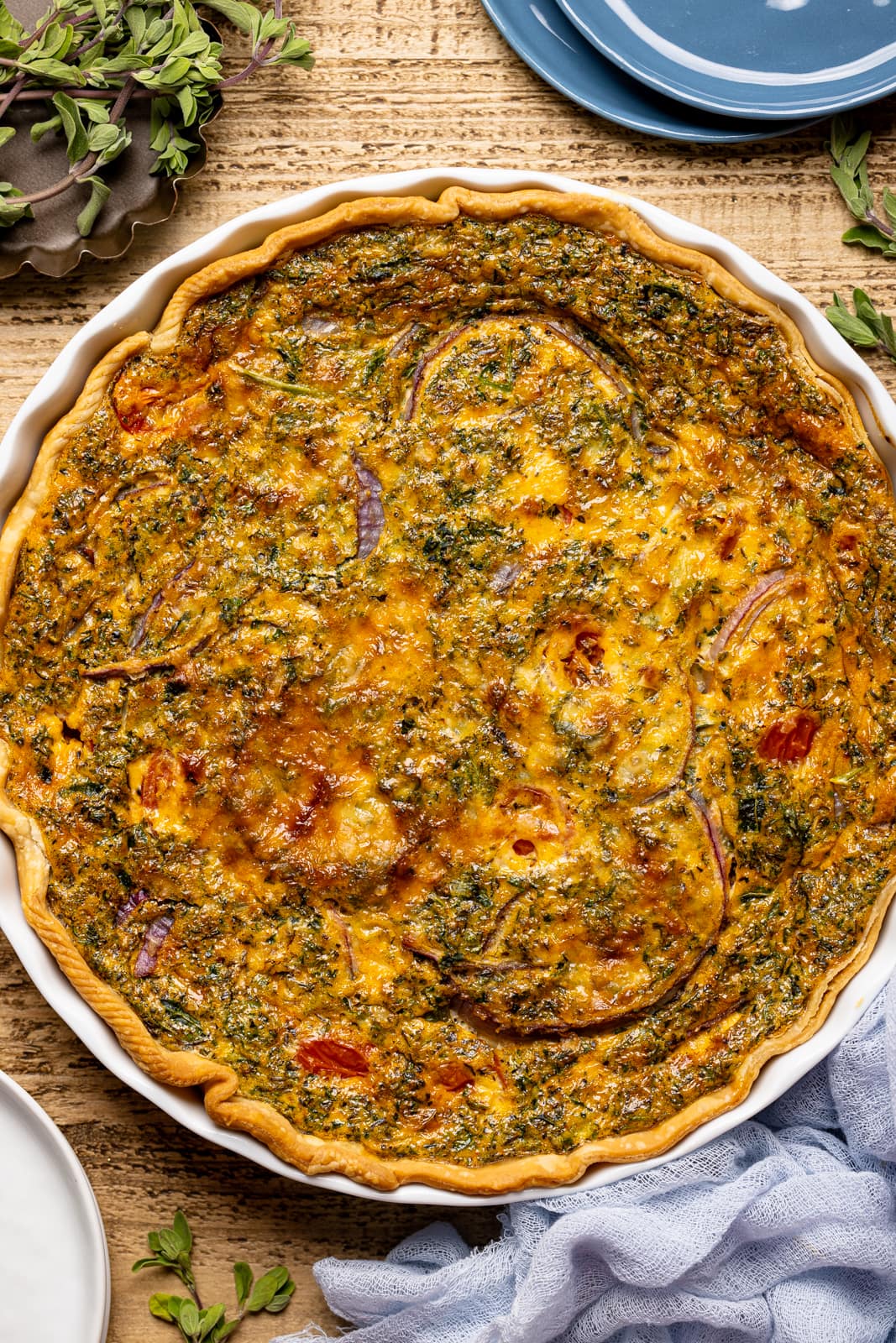 Up close shot of quiche in a baking dish on brown wood table with herbs + seasonings. 