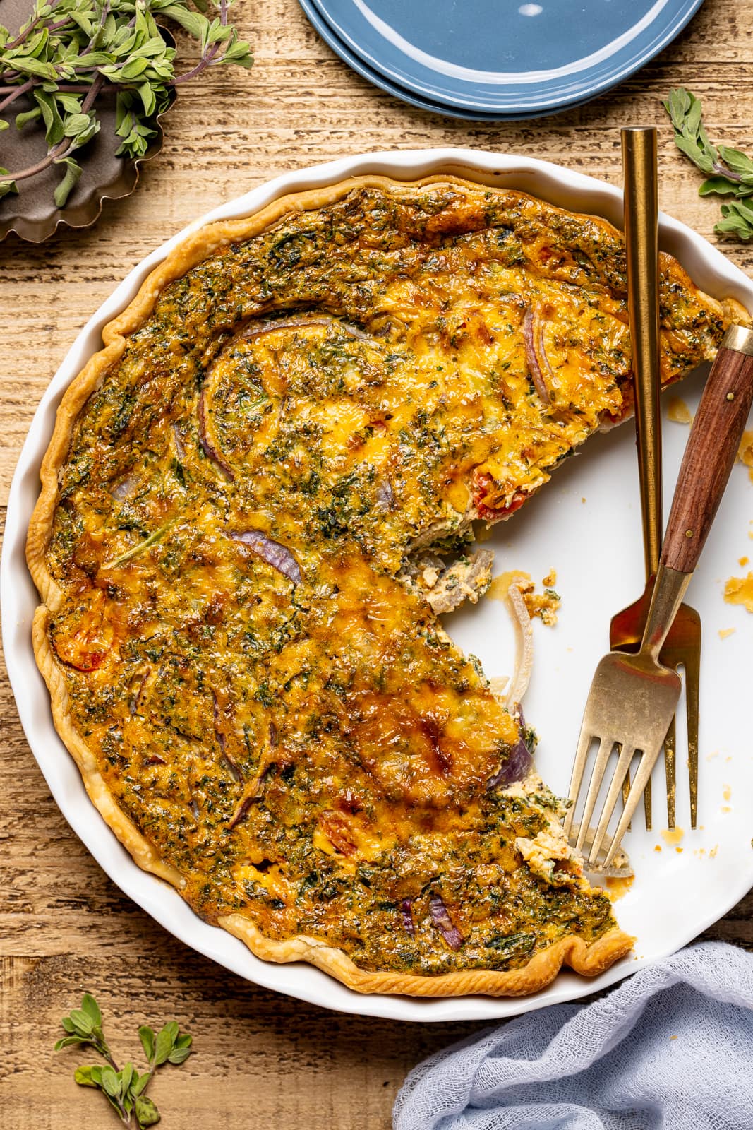 Baked quiche with slices removed on a brown wood table with two forks and a blue napkin and herbs and seasonings. 