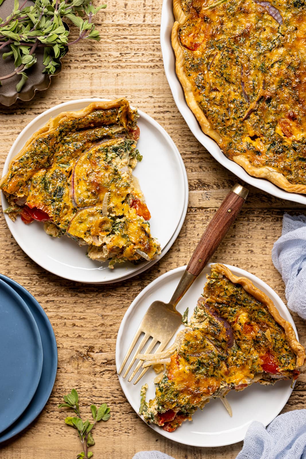 Two slices of quiche on seperate white plates on a brown wood table with blue plates and napkins. 