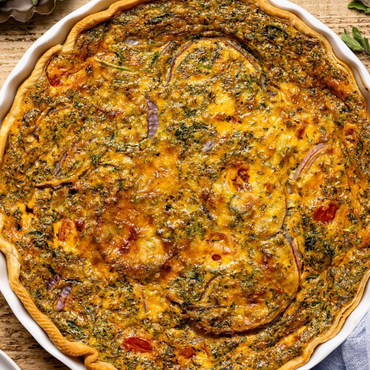 Up close shot of quiche in a baking dish on brown wood table with herbs + seasonings.