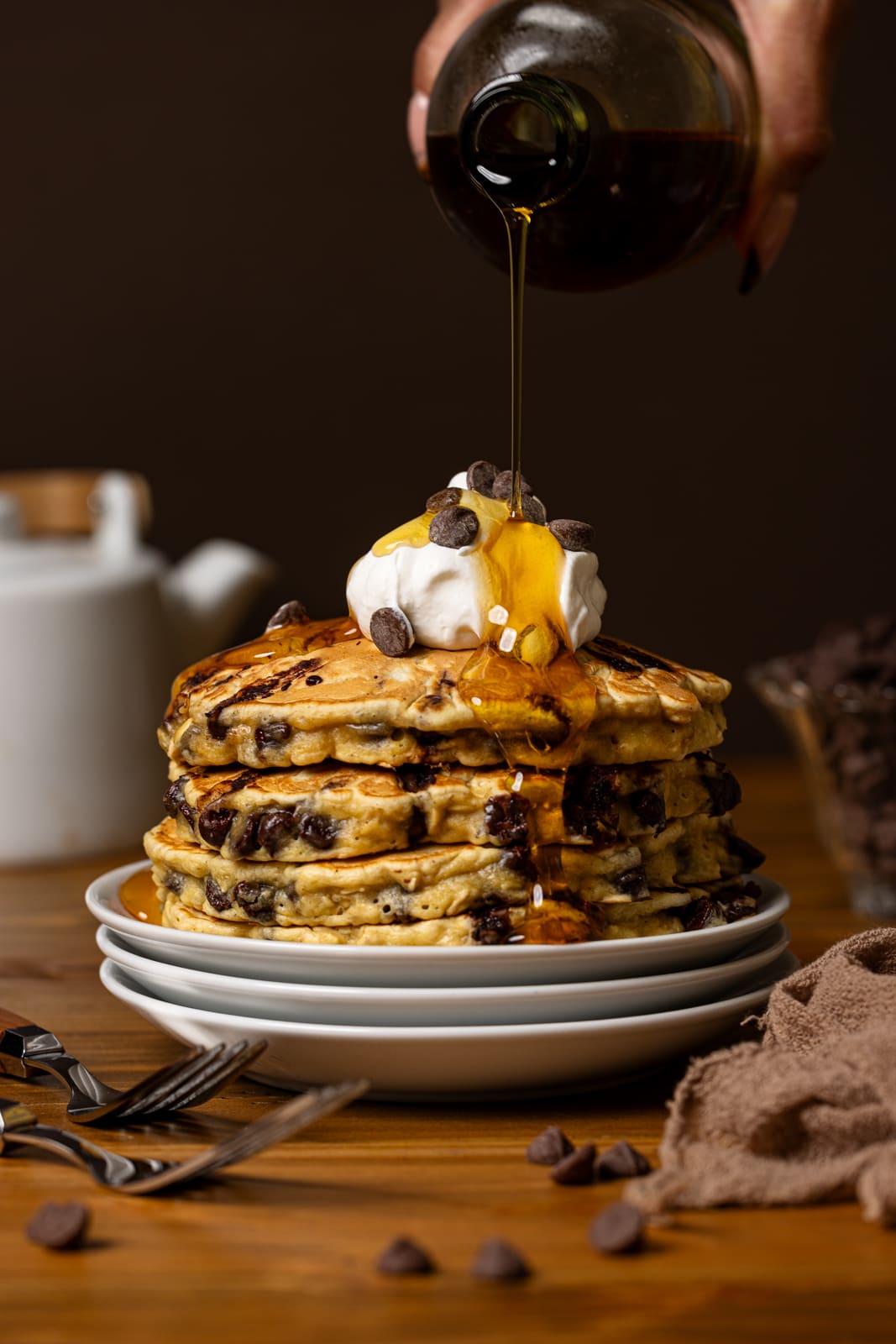 Syrup being poured on a stack of pancakes with whipped cream and chocolate chips.
