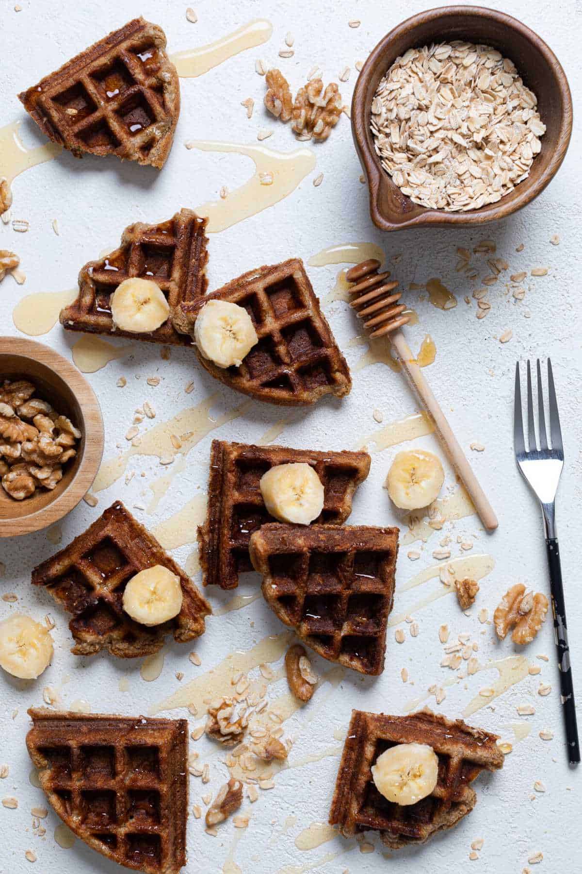 Pieces of Flourless Vegan Banana Oats Waffles on a countertop with honey, bananas, nuts, oats, and utensils.