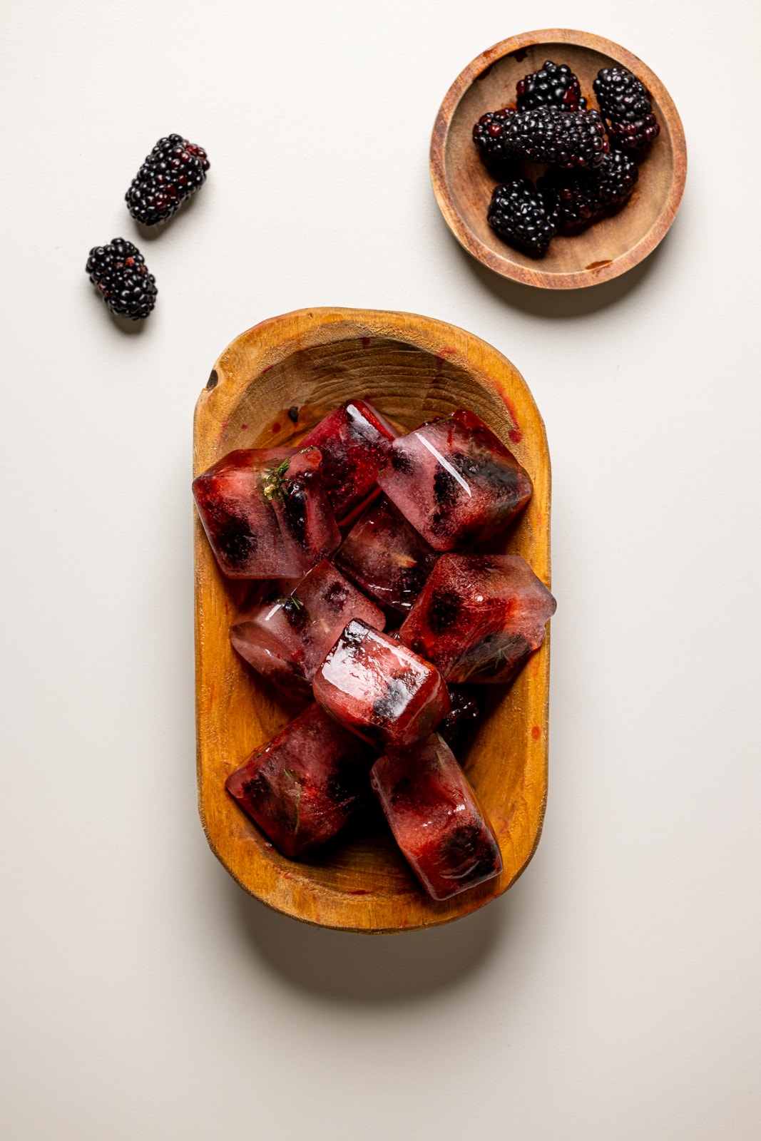 Ice cubes with blackberries in a brown wood bowl on a light grey table.