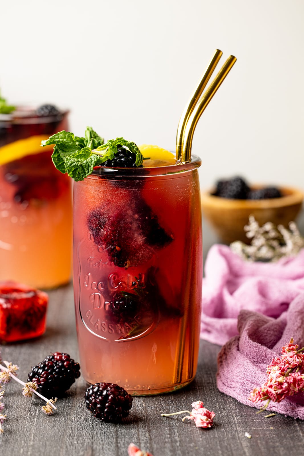 Iced tea in two large jars on a grey wood table with two gold straws with blackberries and lemons.