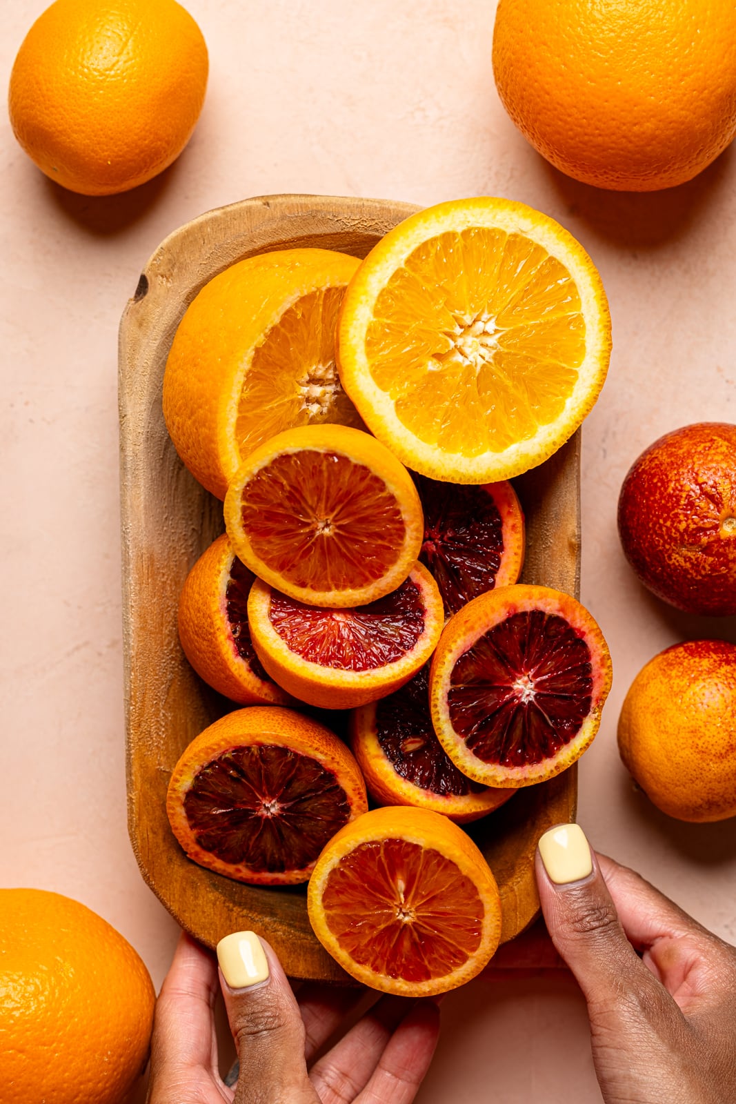 A wooden bowl full of blood oranges being held on a peach colored table.