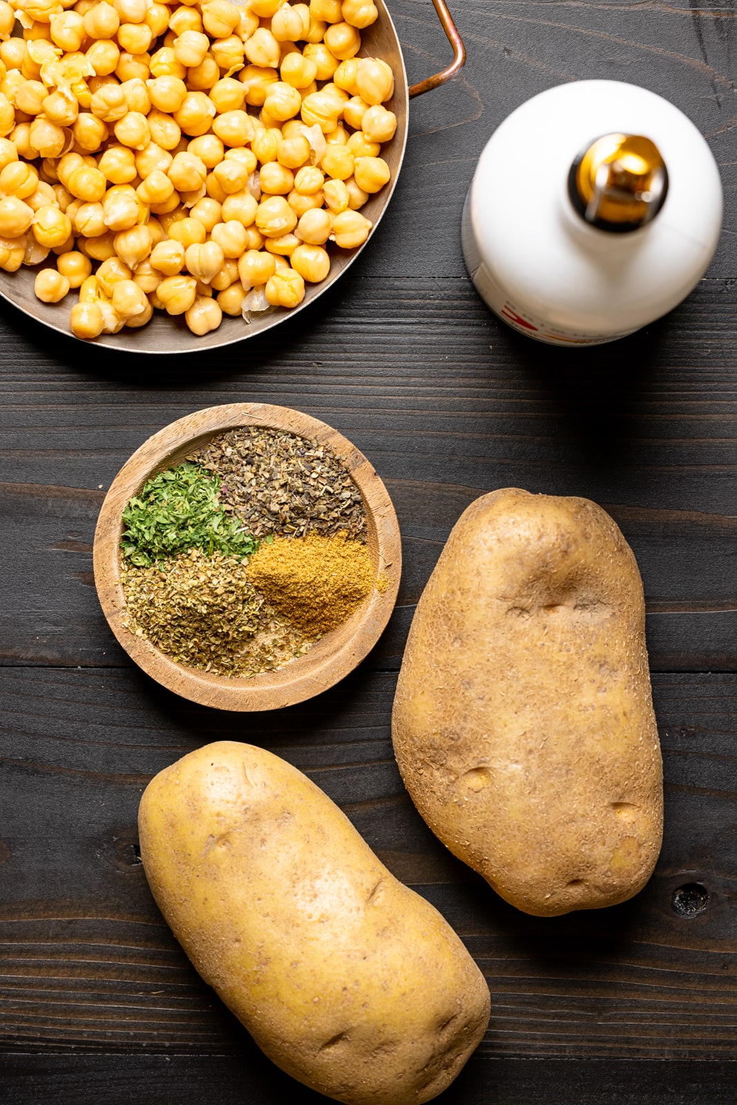 Ingredients on a black wood table including potatoes, olive oil, chickpeas, and herbs and seasonings.