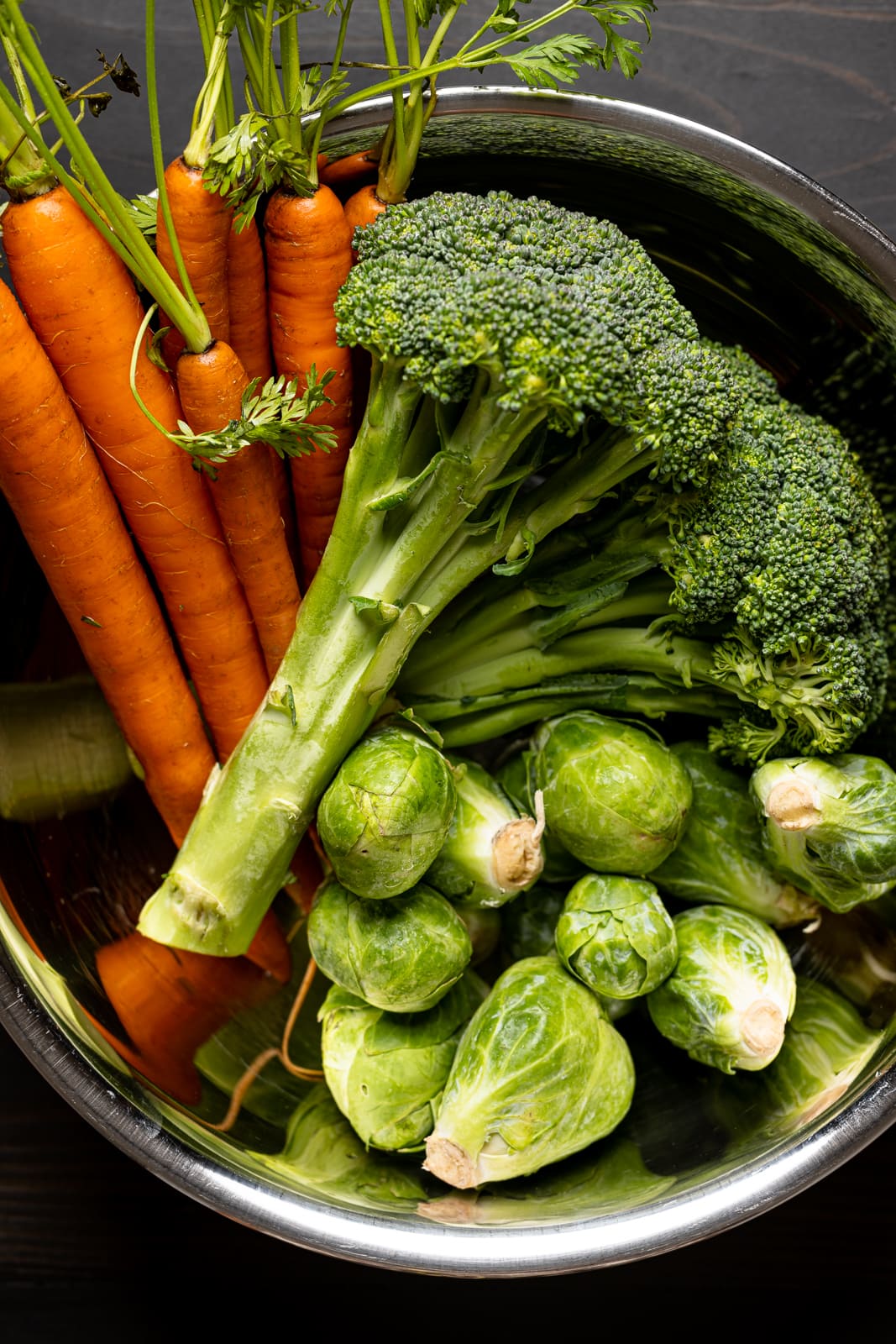 Broccoli, brussels sprouts, and carrots in a large silver bowl on a black table.