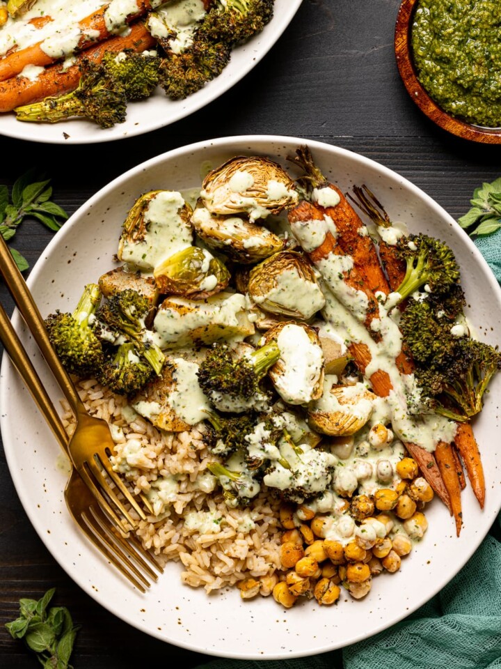 Veggies in two low bowls on a black table with pesto sauce and two forks and herbs.