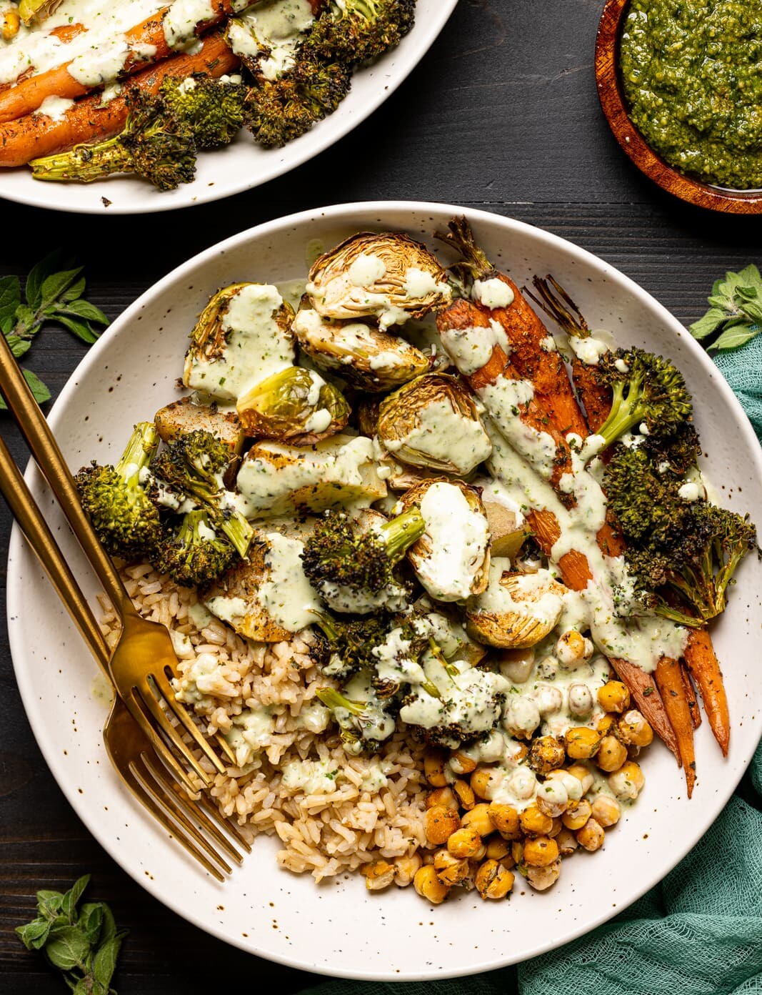 Veggie bowl in two low bowls on a black table with pesto sauce and two forks and herbs.