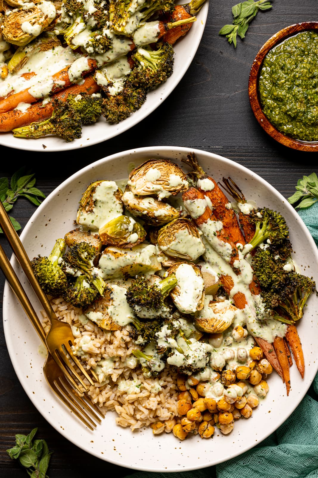 Veggies in two low bowls on a black table with pesto sauce and two forks and herbs.