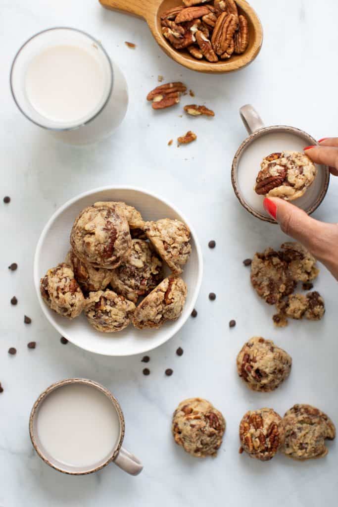 Woman dipping a Dairy-Free Chocolate Chip Pecan Cookie into milk next to a bowl of cookies.