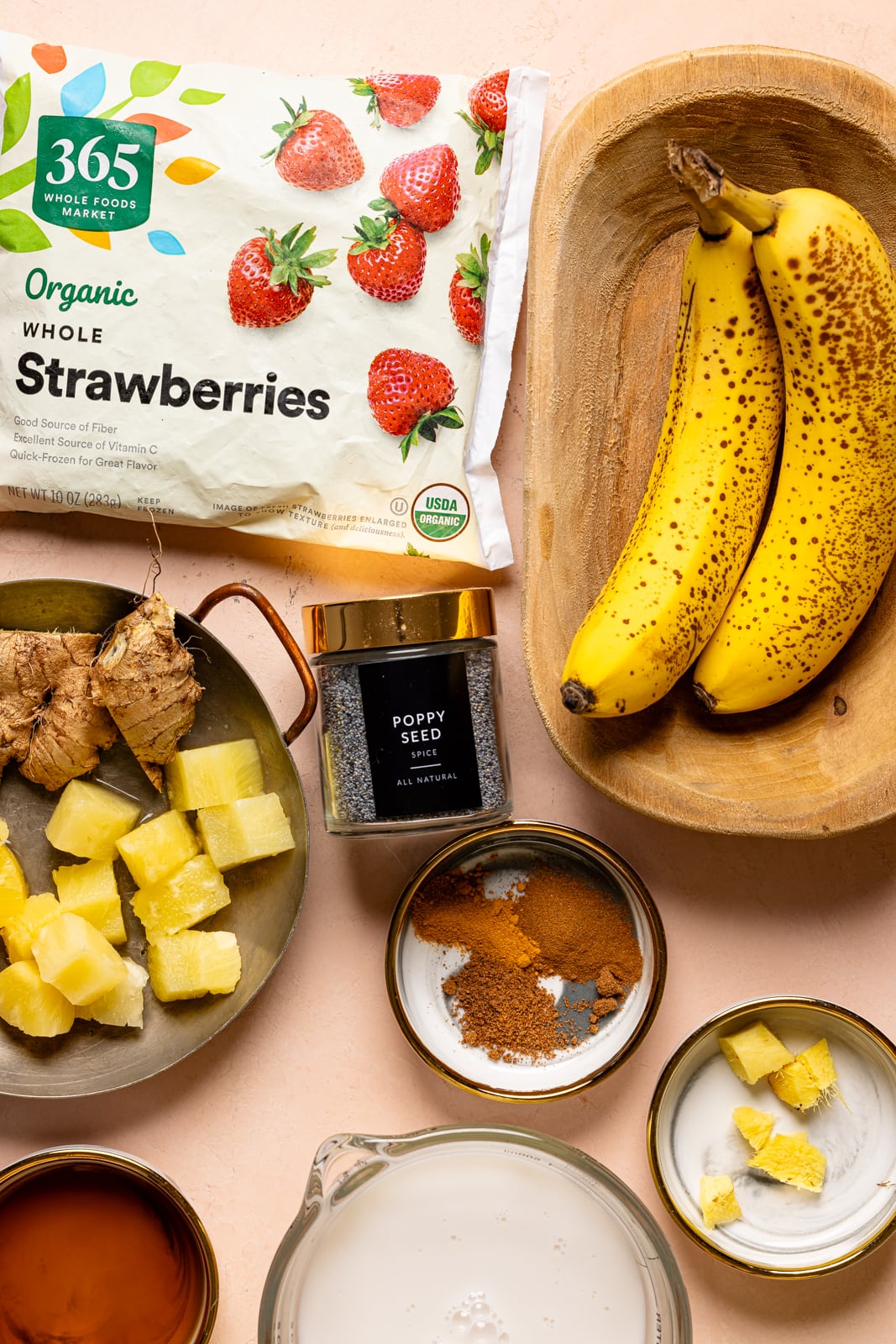 Ingredients on a peach table including frozen strawberries, bananas, pineapples, ginger, spices, milk, and maple syrup.