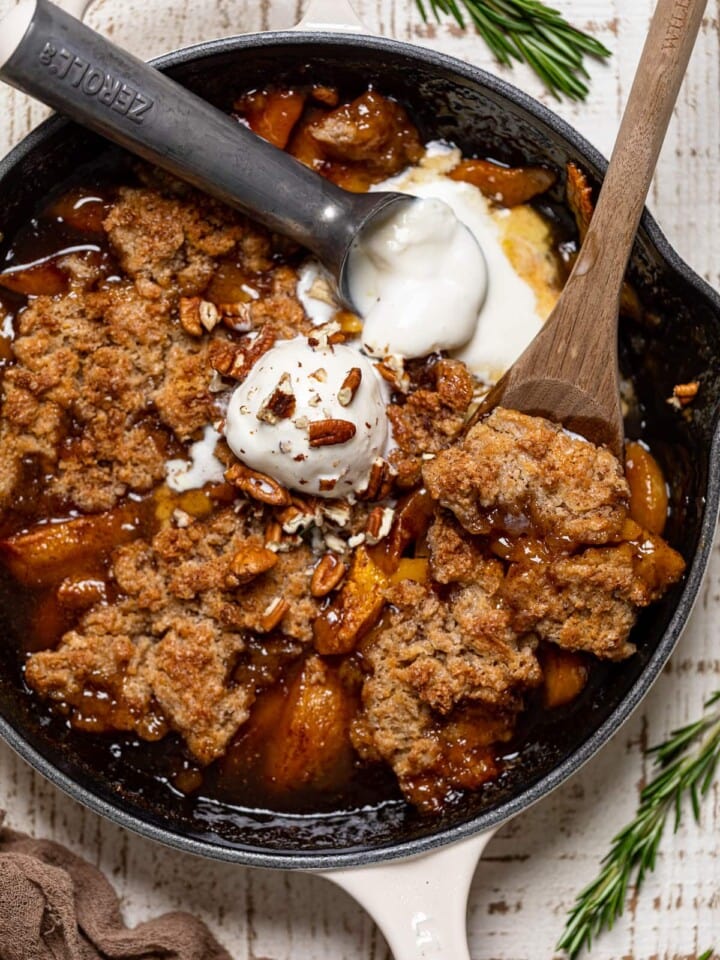 Wooden spoon and ice cream scoop in a skillet of Vegan Southern Peach Cobbler.