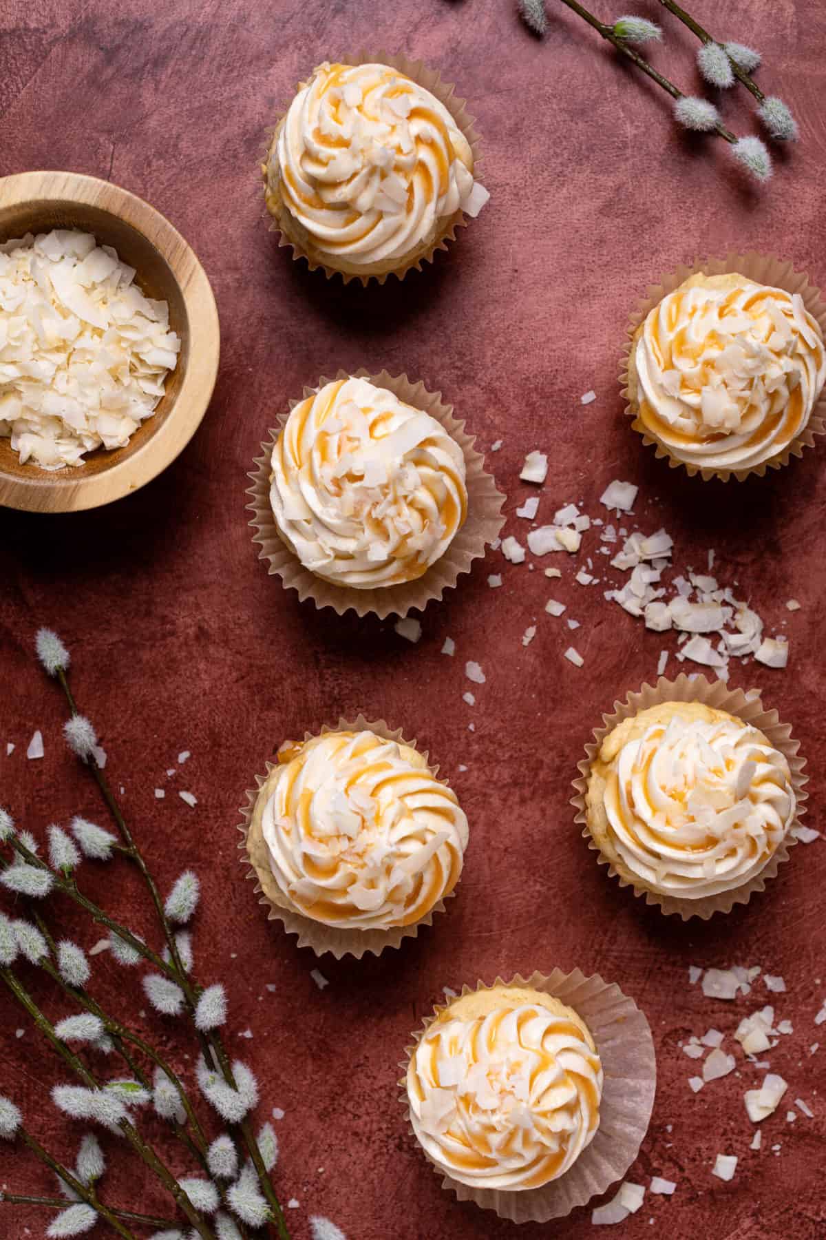 Easy Caramel Coconut Cupcakes in paper wrappers on a red surface with coconut flakes.