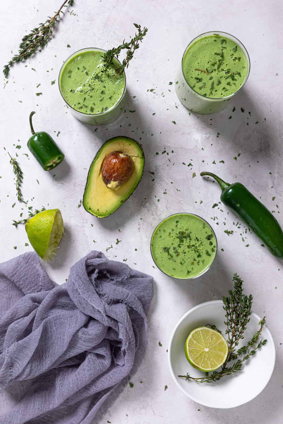 Glasses of Super Green Detox Smoothie on a table with avocado, lime, peppers, and other ingredients.