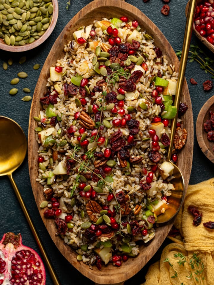 Wild rice in a serving bowl with two gold serving spoons and bowls of ingredients around.