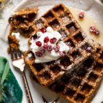 Gluten-free Flourless Gingerbread Waffles topped with whipped cream, pomegranate arils, and powdered sugar.