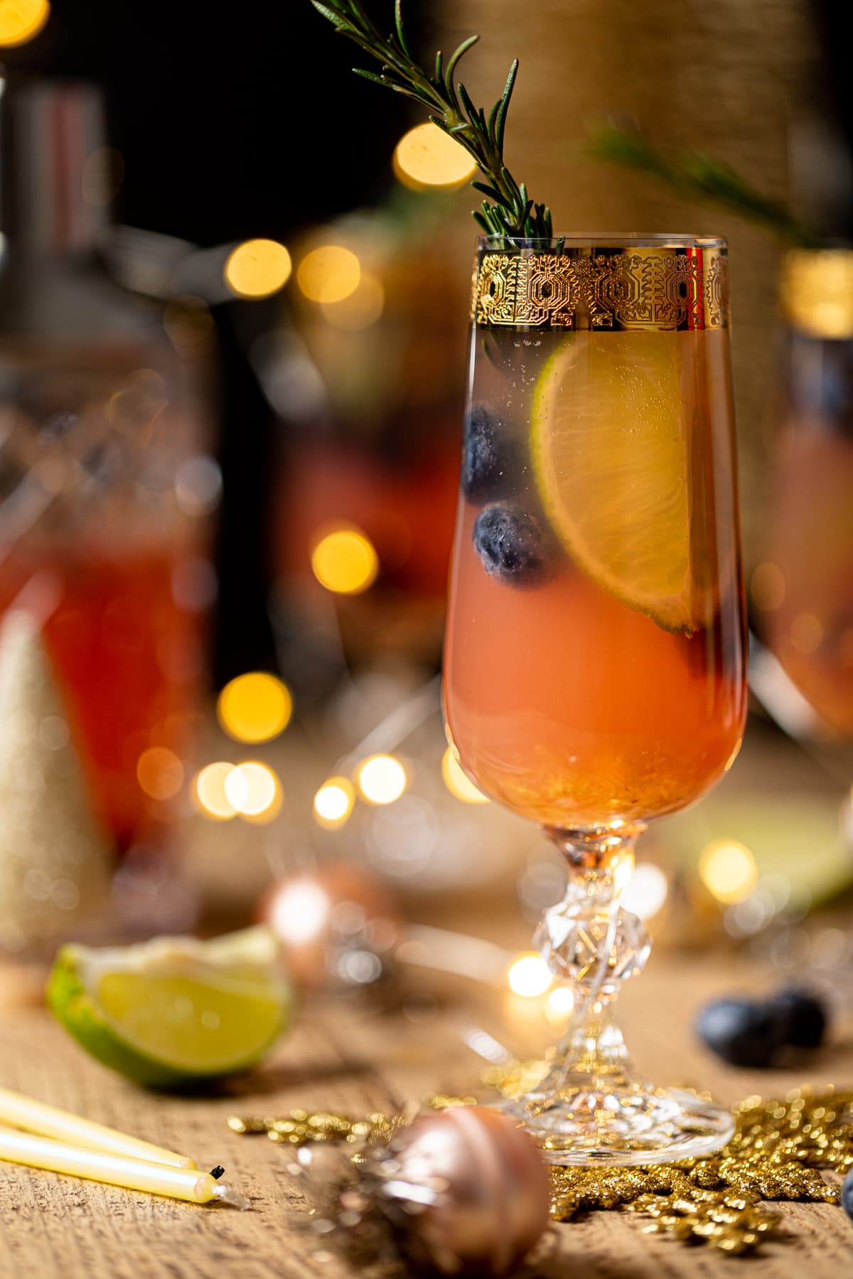 Blueberry Ginger Lime Mocktail topped with a sprig of rosemary in a gold-rimmed, stemmed glass on a table set with Christmas decorations