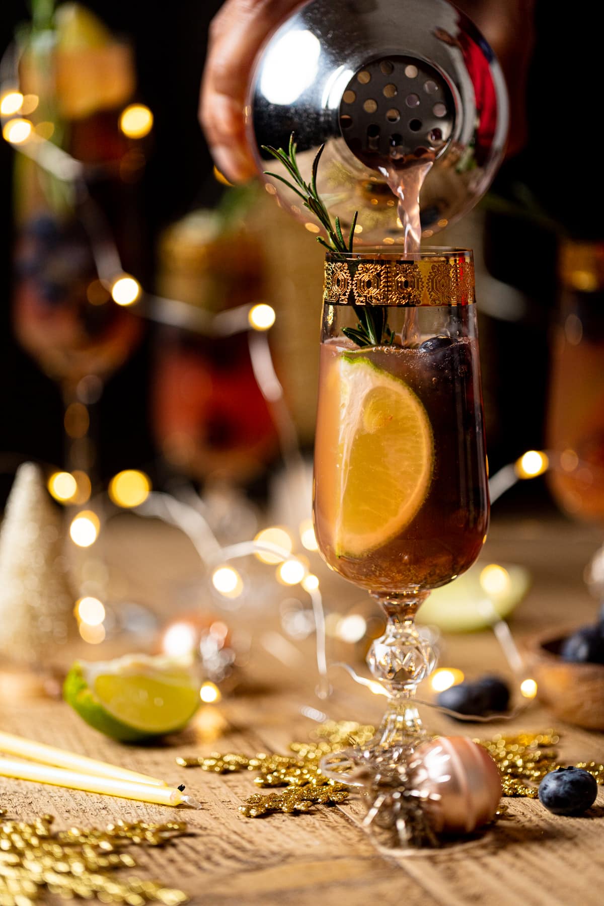 Drink shaker pouring Blueberry Ginger Lime Mocktail into a gold-rimmed, stemmed glass