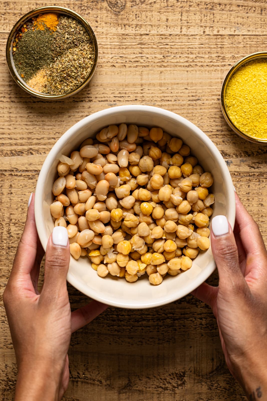 Bowl of chickpeas and white beans being held with hands and herbs + seasonings. 