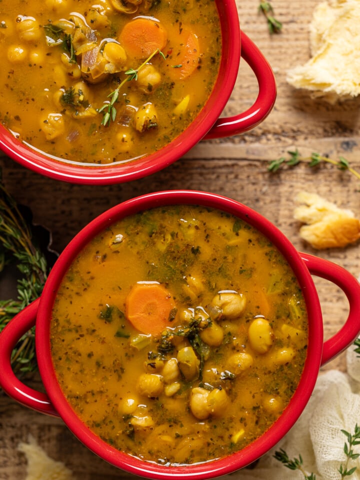 Two red bowls with soup and side of bread on a brown wood table.