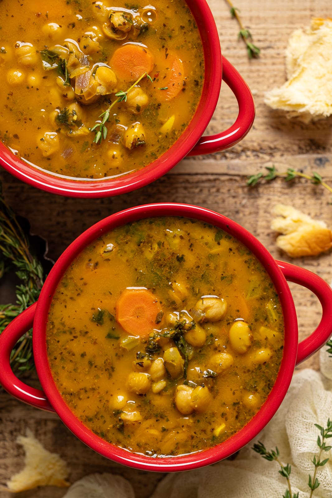 Two red bowls with soup and side of bread on a brown wood table.