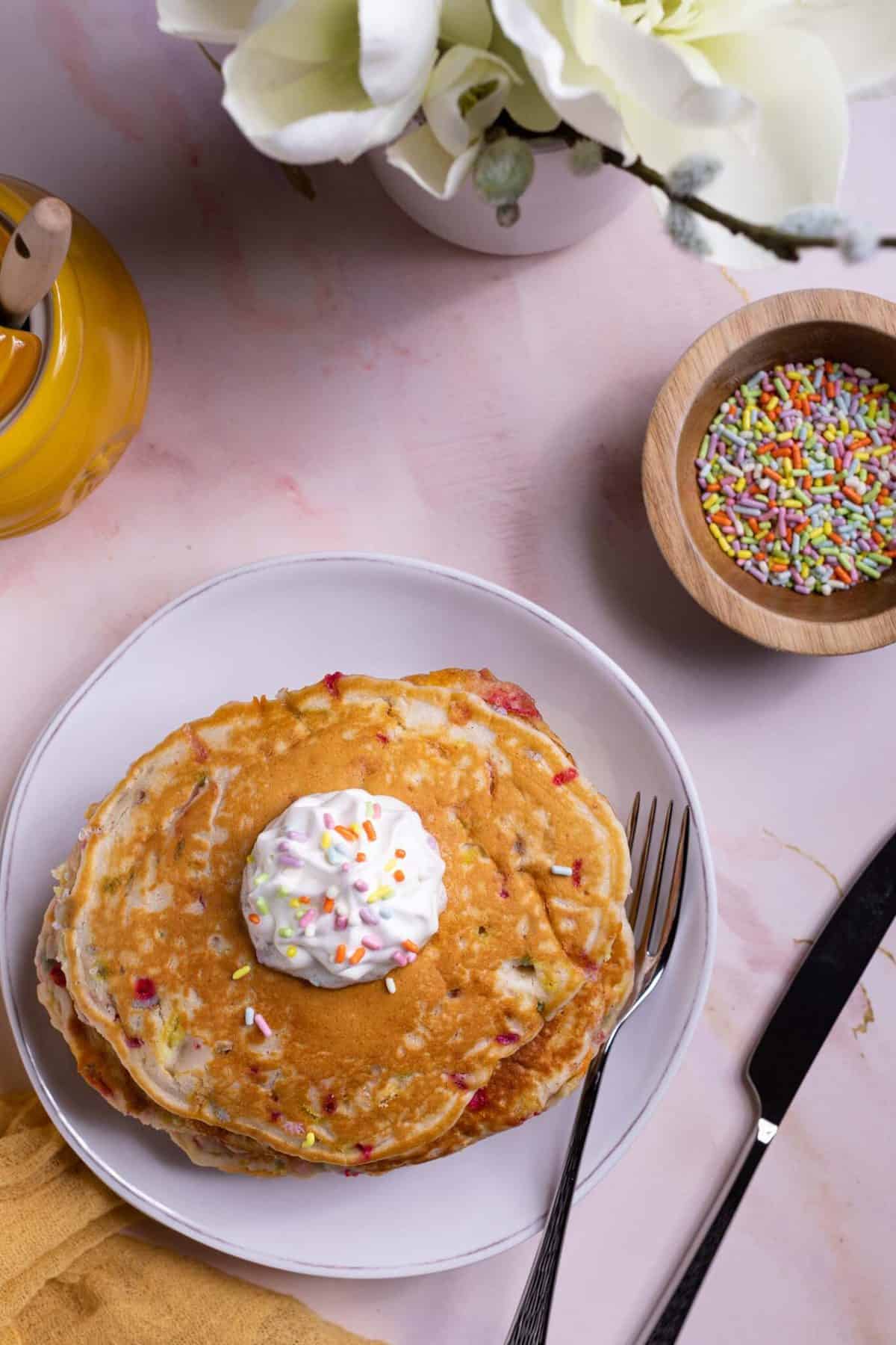 Funfetti Vegan Pancakes in a stack with whipped cream on top and a fork and knife.