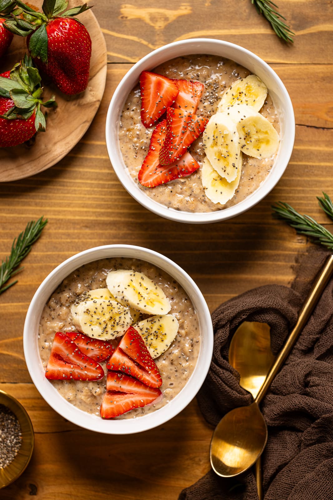 Two bowls of oatmeal with a side of fruit and a spoon on brown wood table.