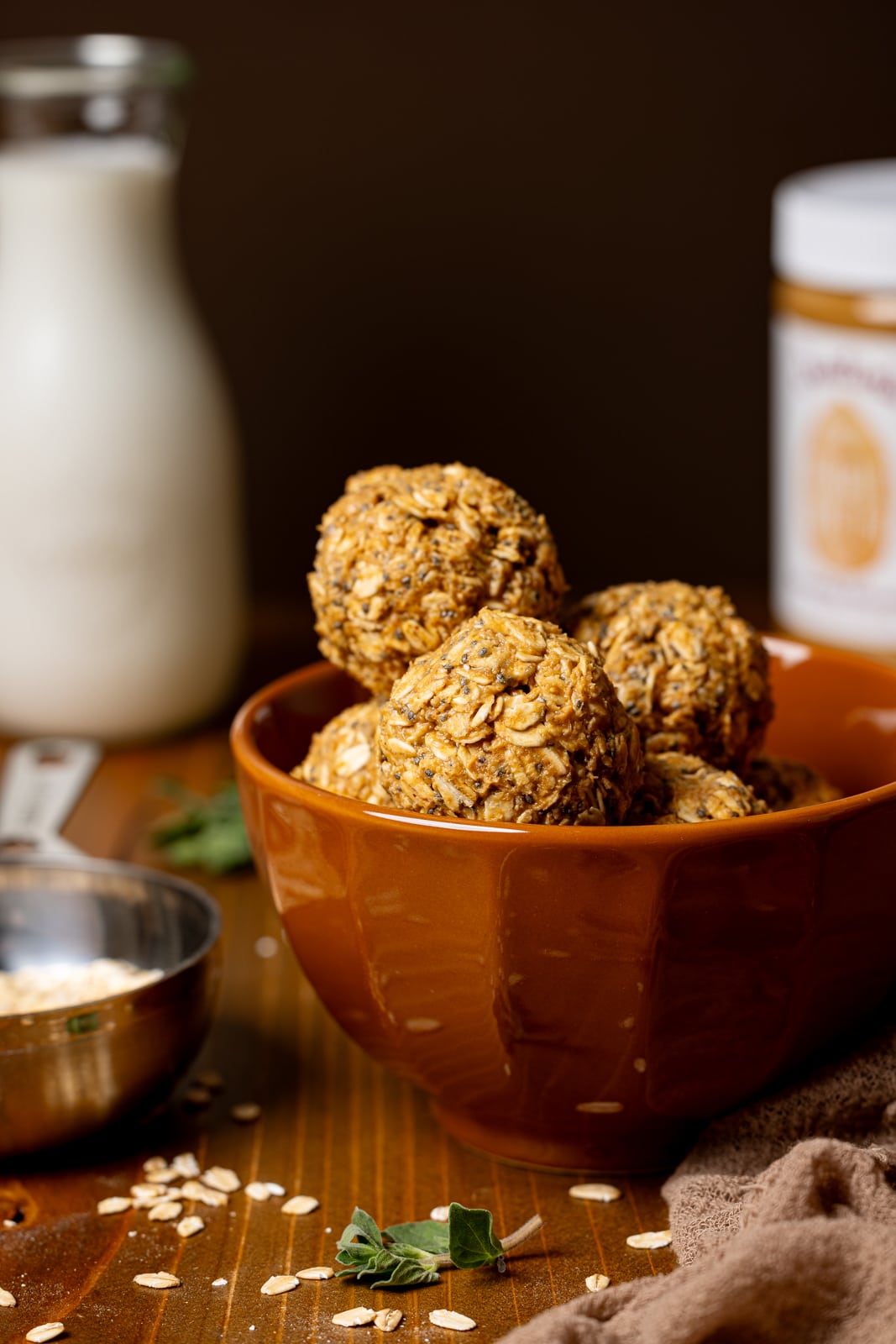 Peanut butter balls in a brown bowl with a measuring cup with oats, glass of milk, and jar of peanut butter.