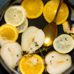 Slices of citrus, pears, and thyme sprigs steeping in a pot with a gold spoon.