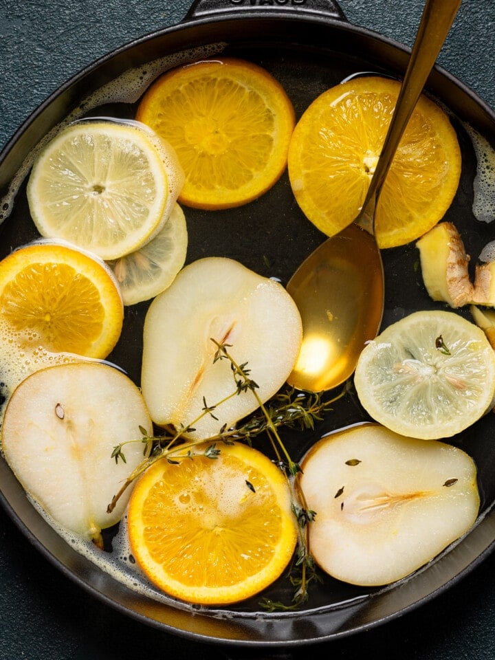Slices of citrus, pears, and thyme sprigs steeping in a pot with a gold spoon.