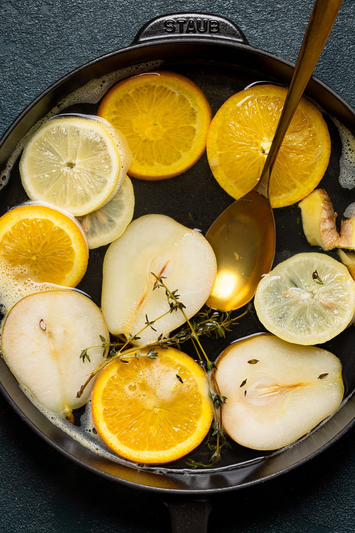 Slices of citrus, pears, and thyme sprigs steeping in a pot with a gold spoon.