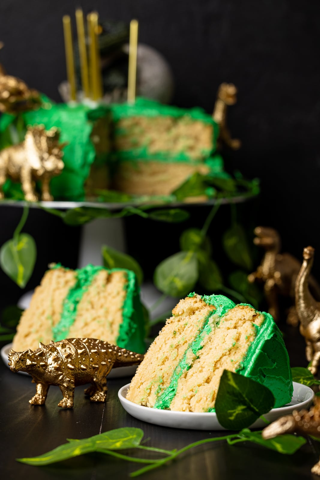 Two slices of cake on a white plate with the whole cake on a cake stand in the black background with gold toy dinosaurs.