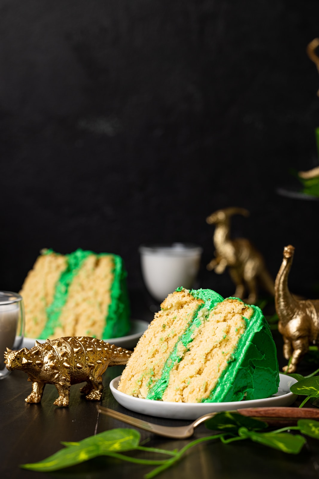 Two slices of cake on white plates with a small glass of milk in the background surrounded by toy dinosaurs.