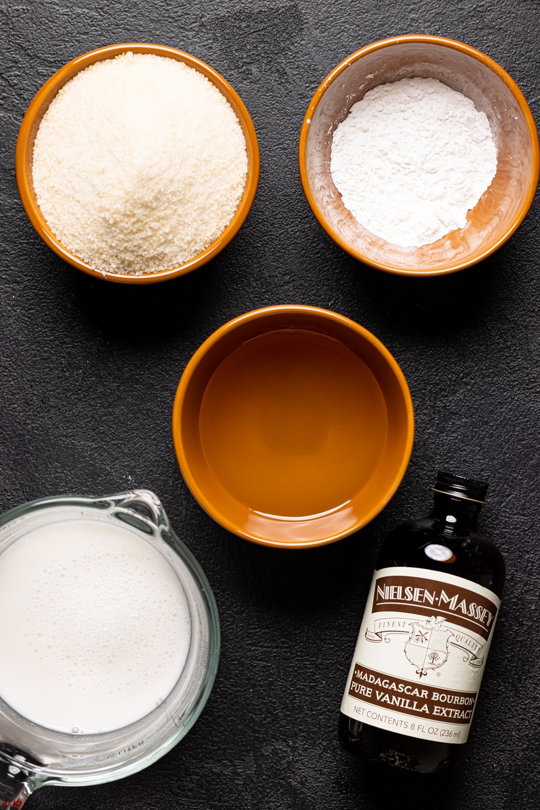 Ingredients on a black table including sugar, vanilla, milk, baking powder, and oil.