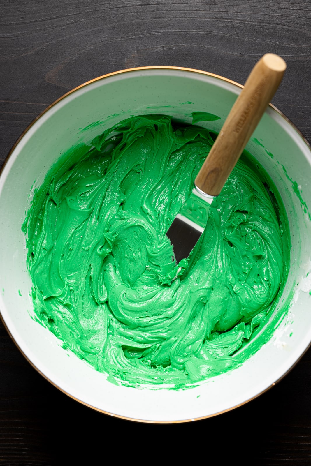 Green colored butter cream frosting in a white bowl on a black table with a cake spatula.