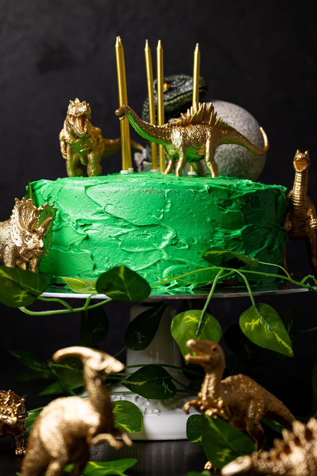 Birthday cake with green frosting and candles with toy dinosaurs on top and surrounding it on a black table.