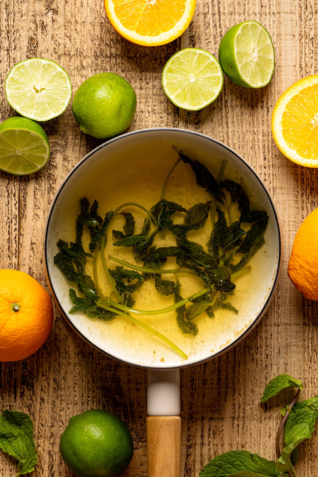 A pot with mint leaves mixture on a brown wood table with slices of orange and lime.