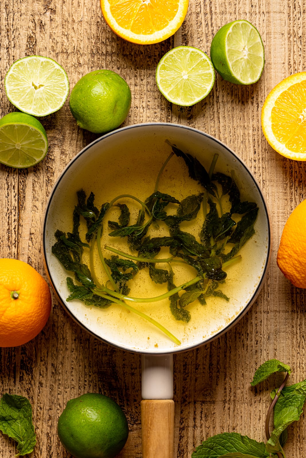 A pot with mint leaves mixture on a brown wood table with slices of orange and lime.