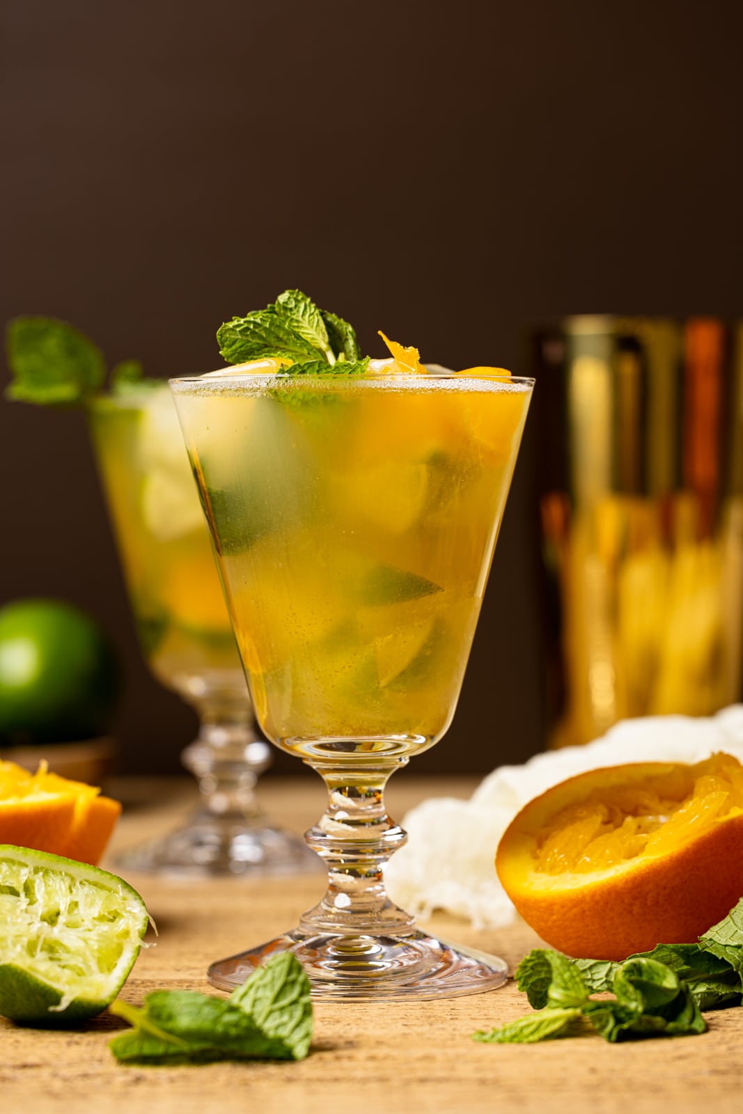 Mocktail in a tall glass on a brown wood table with slices of oranges, lime, mint leaves, with a gold cocktail shaker in the background.