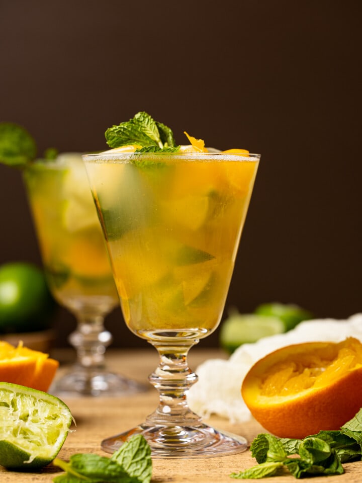 Mocktail in a tall glass on a brown wood table with slices of oranges, lime, mint leaves, with a gold cocktail shaker in the background.