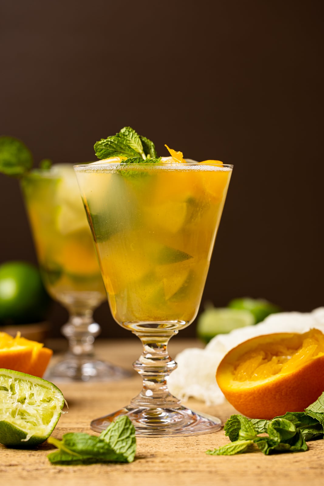 Mocktail in a tall glass on a brown wood table with slices of oranges, lime, mint leaves, with a gold cocktail shaker in the background.